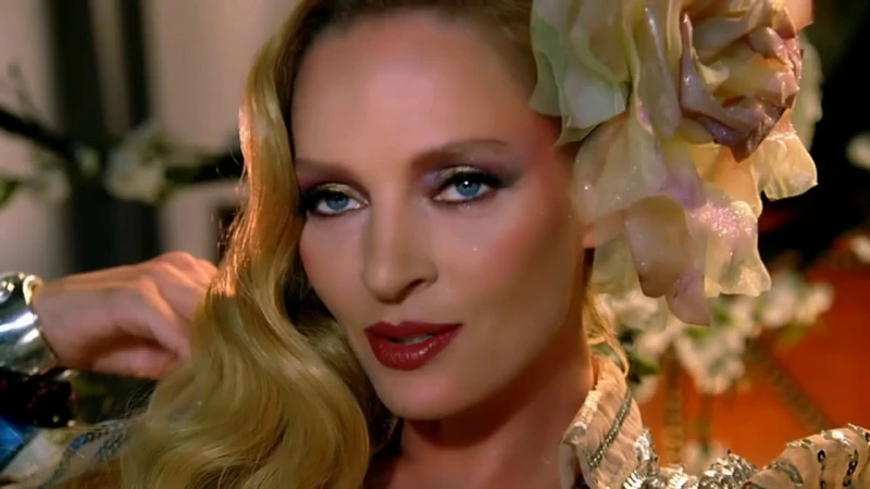 Close-up portrait of Uma Thurman wearing a large floral hairpiece and styled blonde waves, with dramatic makeup.