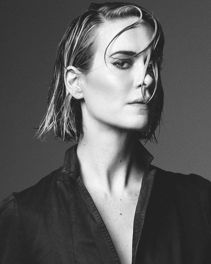 Monochrome portrait of an adult woman with wet blonde hair, wearing a dark collared shirt, against a gray background.