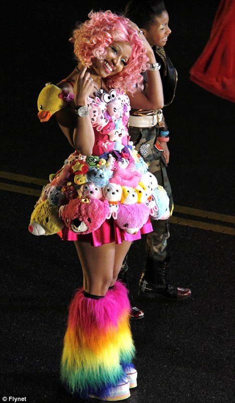 A smiling woman with pink hair in a dress made of stuffed animals and furry rainbow leg warmers posing with another person in the background.