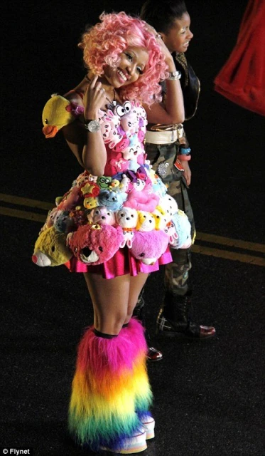 A smiling woman with pink hair in a dress made of stuffed animals and furry rainbow leg warmers posing with another person in the background.