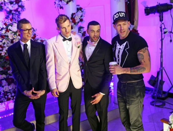 Four adult men dressed in formal wear, including tuxedos and a t-shirt with a tuxedo print, pose in front of a floral decorated backdrop.