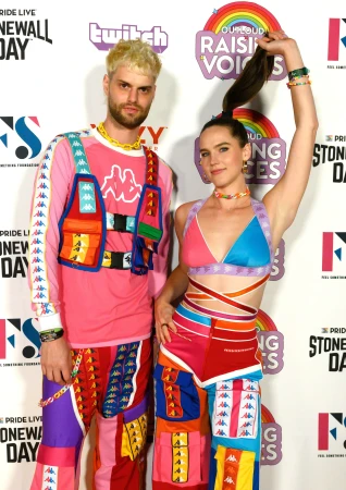 Two people stand in front of a 'Stonewall Day' backdrop. One has blond hair and a pink Kappa outfit with a vest. The other has a ponytail and multicolored Kappa clothing.