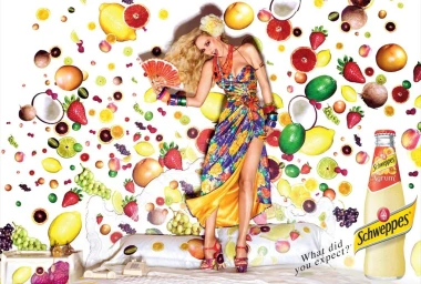 Blonde woman in colourful printed dress holding a fan standing amidst floating fruits and a Schweppes bottle
