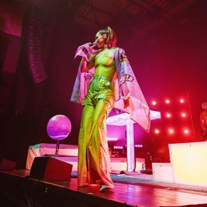 A female singer on stage holding a microphone, wearing lime green cargo pants and a printed cape under dramatic red stage lighting.