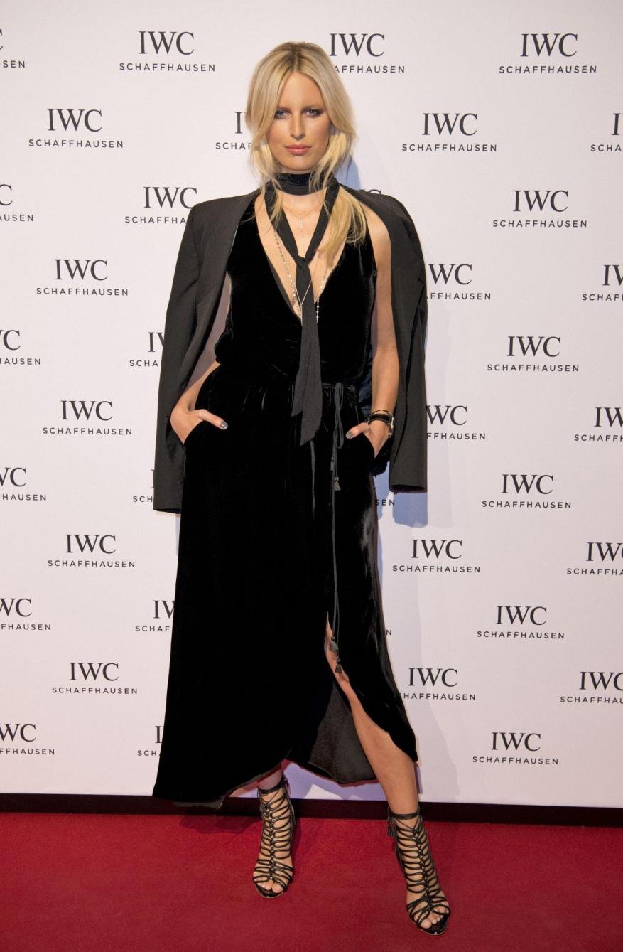Blonde model in black velvet dress and strappy heels poses on a red carpet in front of an IWC Schaffhausen branded backdrop.