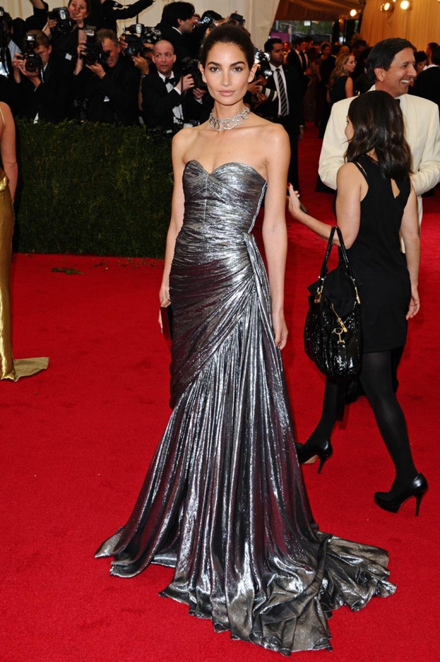Lily Donaldson poses on a red carpet wearing a silver strapless gown and diamond necklace at a premiere event with photographers.