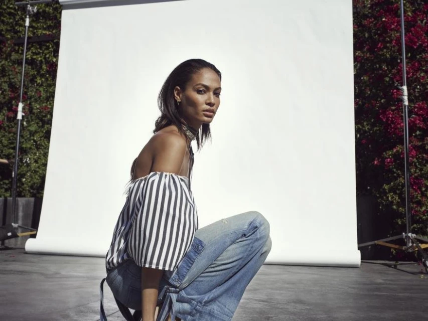 Elegant woman poses crouching in front of a bright white backdrop with light blue jeans and off-the-shoulder striped blouse.