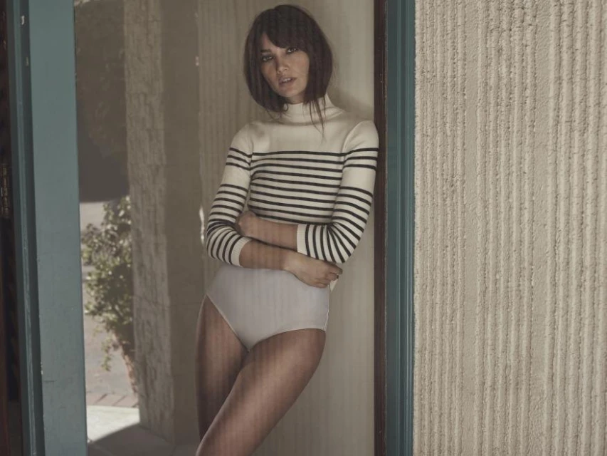 Fashion model in striped turtleneck sweater and briefs poses near a blue-framed window with textured wall in soft, natural light.