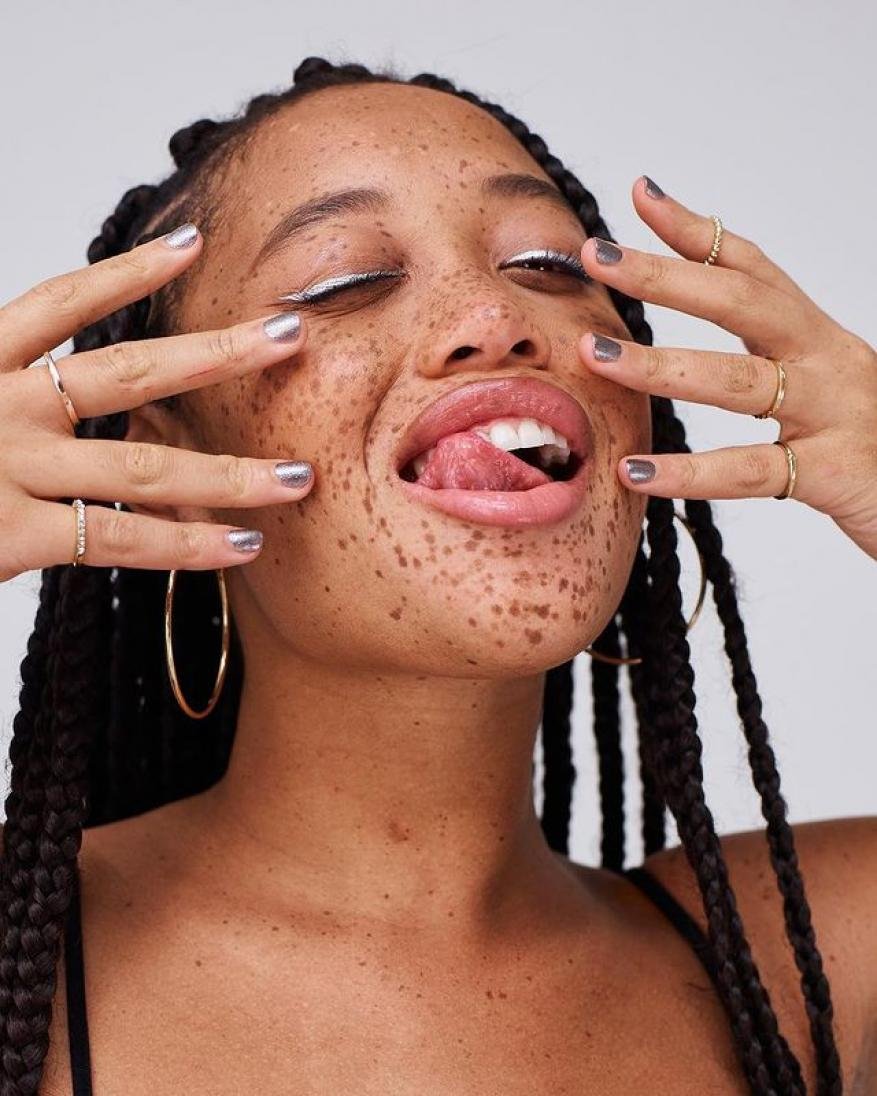 Close-up studio shot of a Black woman with braided hair and freckles, wearing makeup and jewelry while sticking her tongue out.