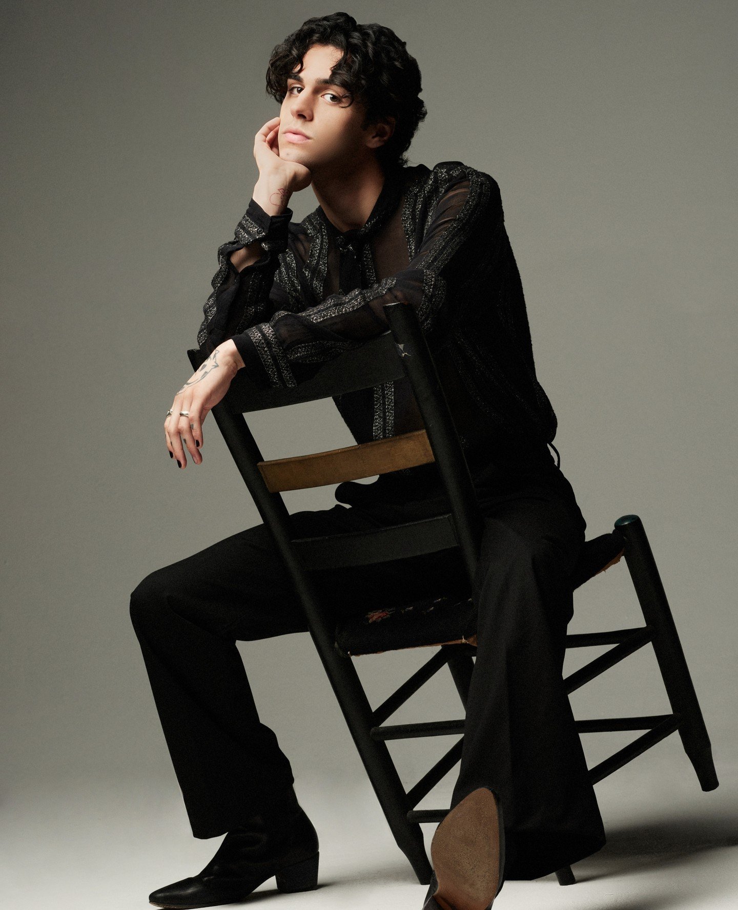 Portrait of a young adult with dark curly hair, wearing a patterned black shirt, sitting on a black chair in a photo studio.