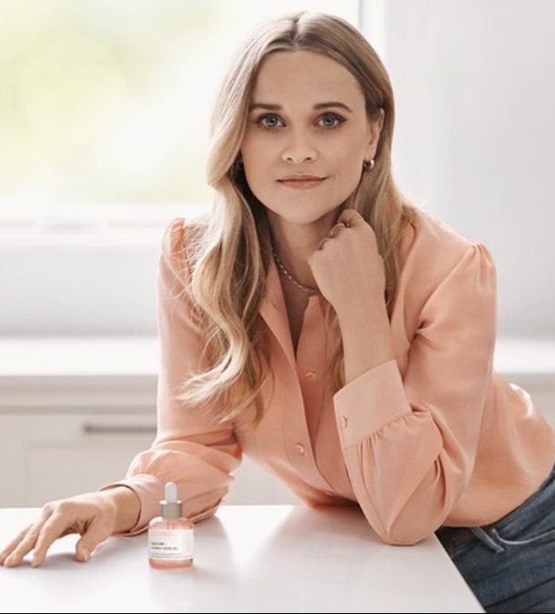 Blonde woman in pink blouse leans on a white table near a window with a bottle of skincare serum in a bright indoor setting.