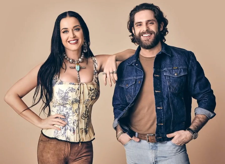 Katy Perry and Thomas Rhett pose in a close-up shot. Perry wears a floral corset and Rhett sports a denim jacket, both smiling warmly.