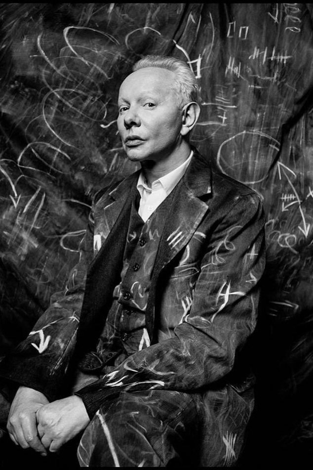 Portrait of a light-skinned adult male in a dark patterned suit with white markings, seated indoors against a textured background with scribbles.