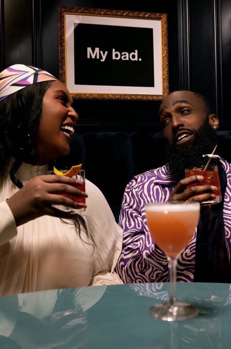Two smiling African-descent adults at a bar with drinks, under framed wall art that reads 'My bad', indoors with dark, upscale decor.