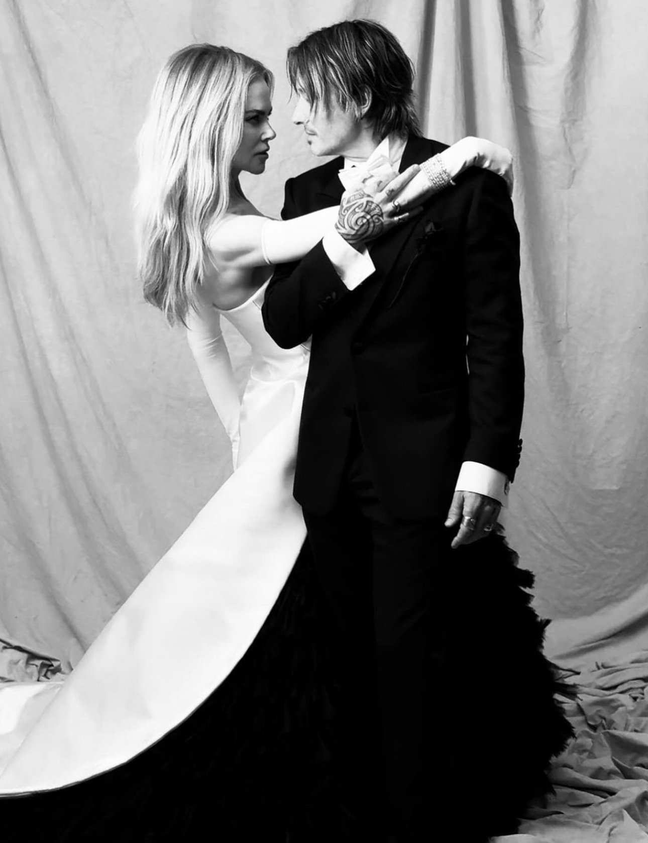 Black and white shot of a blond woman in a white dress and a man in a black suit facing each other with arms around one another.