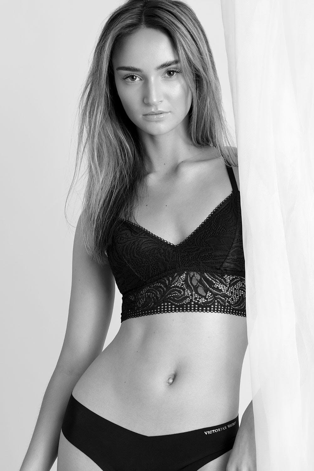 Black and white portrait of a young woman posing in lingerie with a neutral expression against a white backdrop with soft lighting.