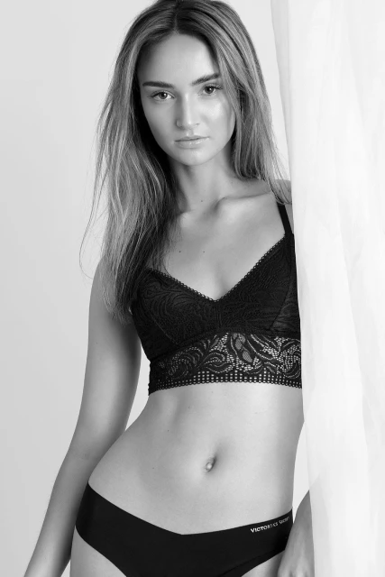 Black and white portrait of a young woman posing in lingerie with a neutral expression against a white backdrop with soft lighting.