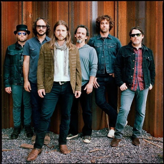 Group portrait of six adult men posing against a textured rusted metal wall, wearing casual clothing, in an outdoor setting.