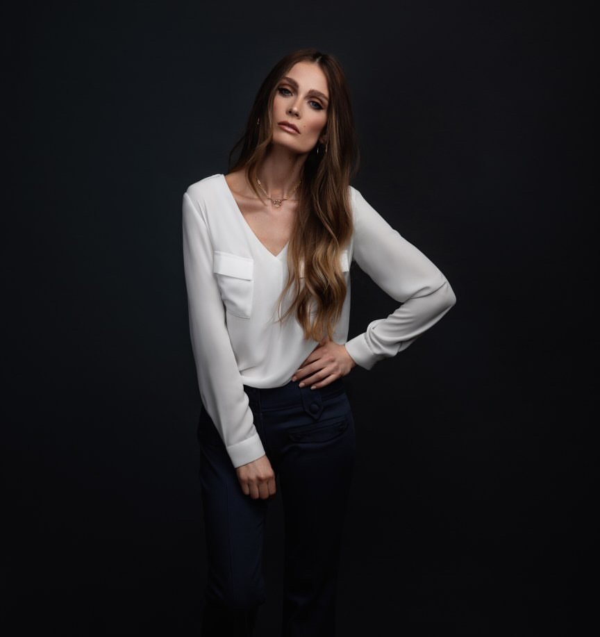 Full shot of a woman in a white blouse and dark pants posing against a dark background in a portrait orientation with indoor lighting.
