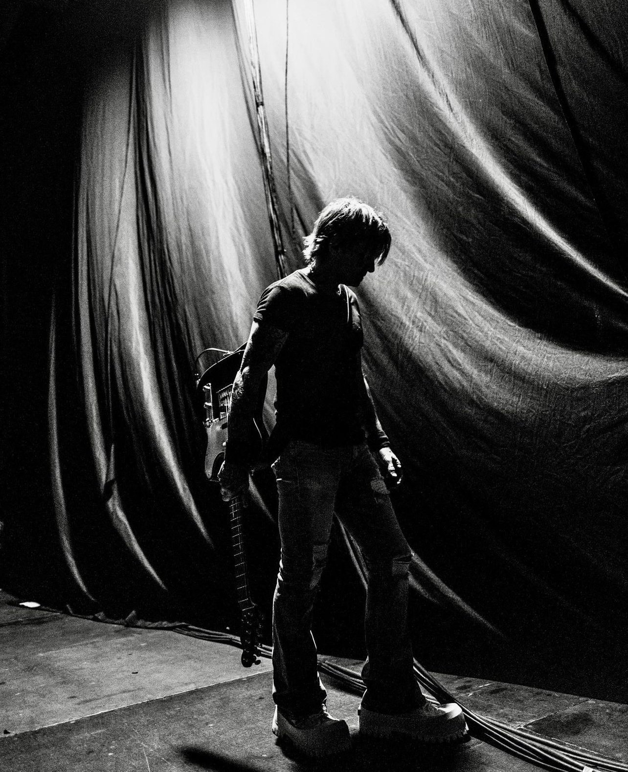 Black and white portrait of a musician holding a guitar, standing on stage in front of black curtains, illuminated by stage lighting.