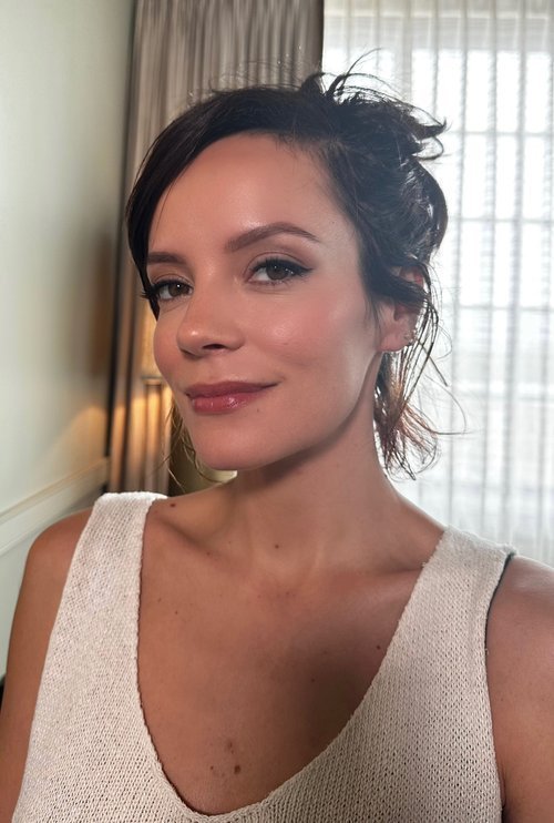 Close-up of a woman with brown hair, wearing a cream-colored sleeveless top, posing indoors with natural lighting near a curtained window.