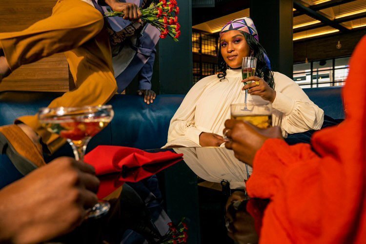 Woman in cream dress sits at a table in an indoor setting, holding a champagne flute, with people and drinks surrounding the table.