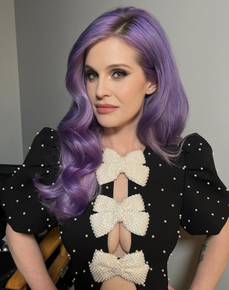 Portrait of a woman with purple wavy hair wearing a black top adorned with pearl white bows and pearl embellishments, smiling slightly.