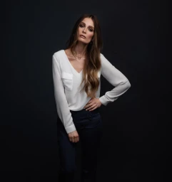 Portrait of a woman wearing a white V-neck blouse and dark pants against a dark background, styled by Katie Galliher.