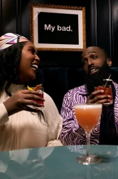 A Black woman and a Black man holding cocktails and laughing, seated at a bar with a framed sign reading "My bad." on the dark wall.