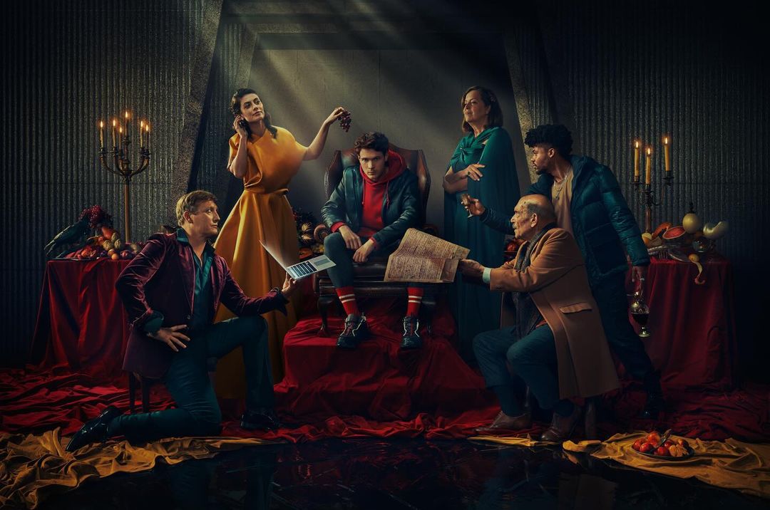 Group of six adults in a dimly lit room with fruit, books, and a laptop, evoking a dramatic, theatrical atmosphere.