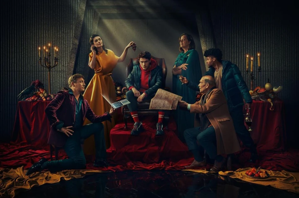 Group of six adults in a dimly lit room with fruit, books, and a laptop, evoking a dramatic, theatrical atmosphere.