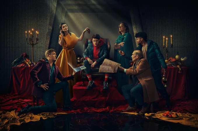 Group of six adults in a dimly lit room with fruit, books, and a laptop, evoking a dramatic, theatrical atmosphere.