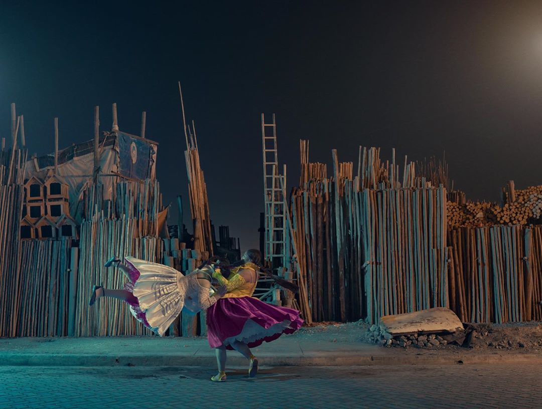 Two dancers in colorful costumes perform in an outdoor urban setting at night, with an artistic backdrop of stacked lumber.