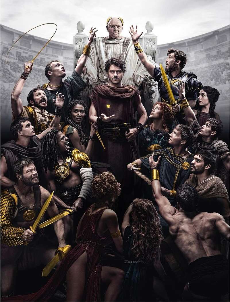 Group of people in ancient Roman attire, some holding swords and other props, gathered around a man in a tunic, with crowd scene backdrop.