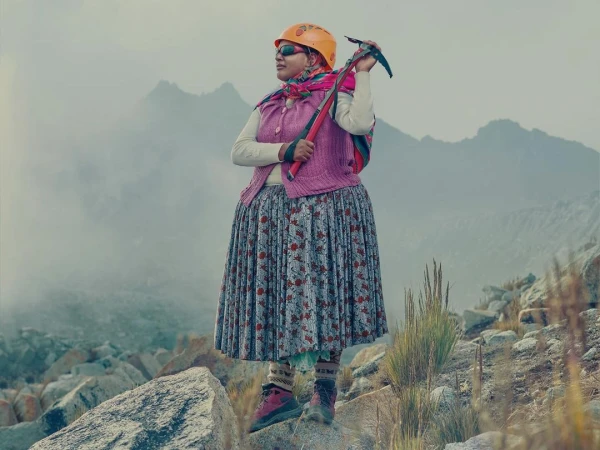 Bolivian woman Cholita climber in traditional dress stands atop a rocky mountain wearing helmet and sunglasses, holding an ice axe.
