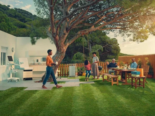 Outdoor family gathering on lawn near modern house; multigenerational group at picnic table, child swings, adult walks with drink.