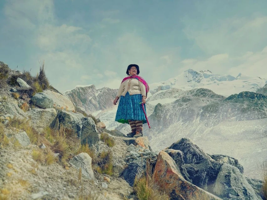 Elderly indigenous woman in traditional clothing stands on rocky mountain terrain with snow-capped peaks, under a cloudy sky in a medium shot.