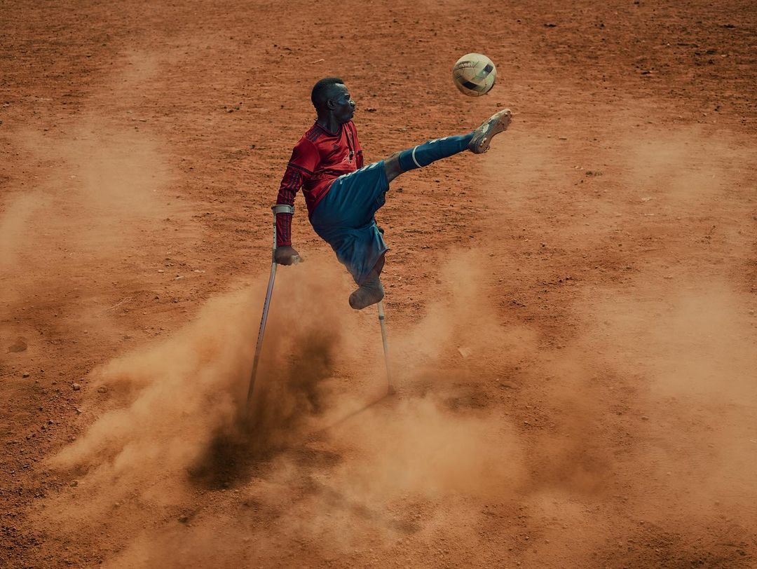 Disabled athlete kicks a soccer ball with a leg supported by crutches on a dusty outdoor field in a powerful display of athleticism.