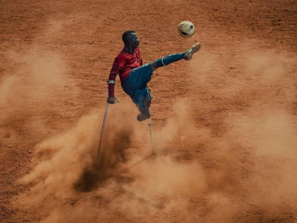 Disabled athlete kicks a soccer ball with a leg supported by crutches on a dusty outdoor field in a powerful display of athleticism.