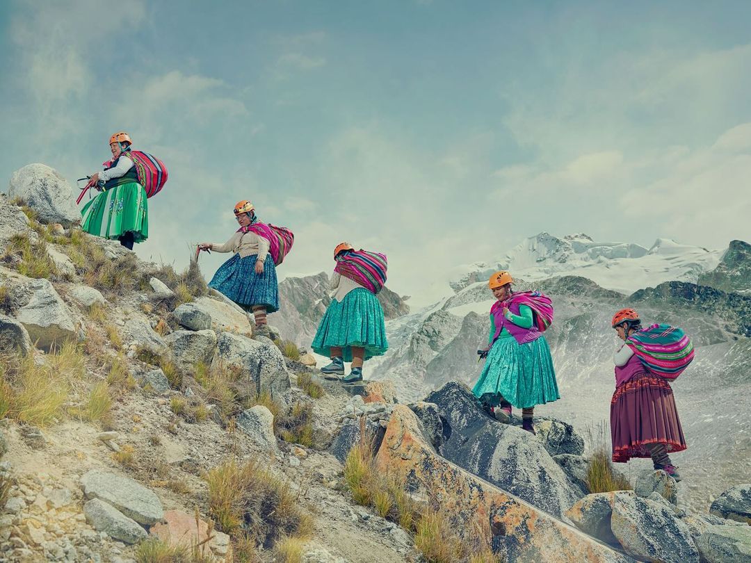 Five women in colorful traditional clothing and helmets climb a rocky mountain, with snow-capped peaks in the distance, on a cloudy day.