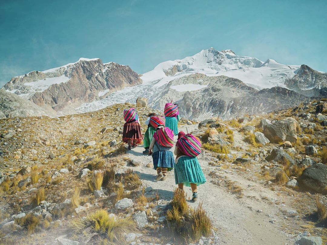 Indigenous women hike toward snow-capped mountain with colorful bundles on their backs. Travel photography showcases local culture.