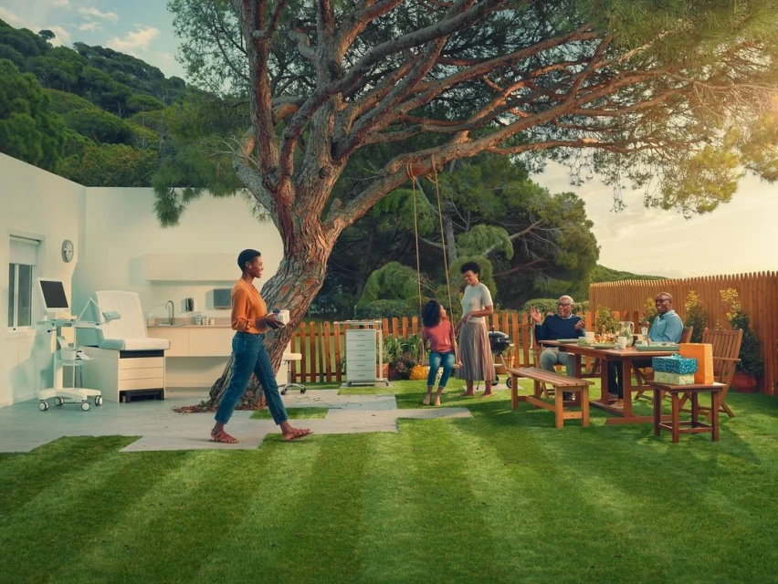 Multi-generational family enjoying an outdoor meal in a bright backyard, with a swing and medical equipment visible near a modern house.