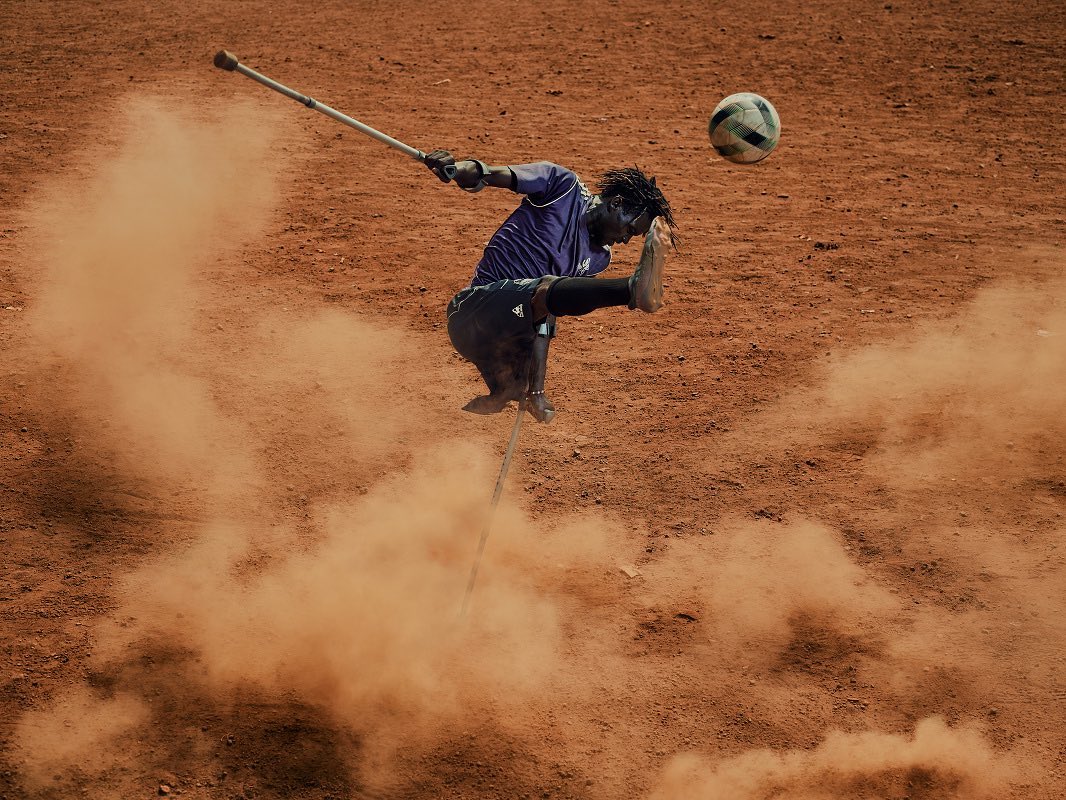 A man with an amputated leg uses crutches to kick a soccer ball in the air on a dusty, reddish-brown surface outdoors on a sunny day.