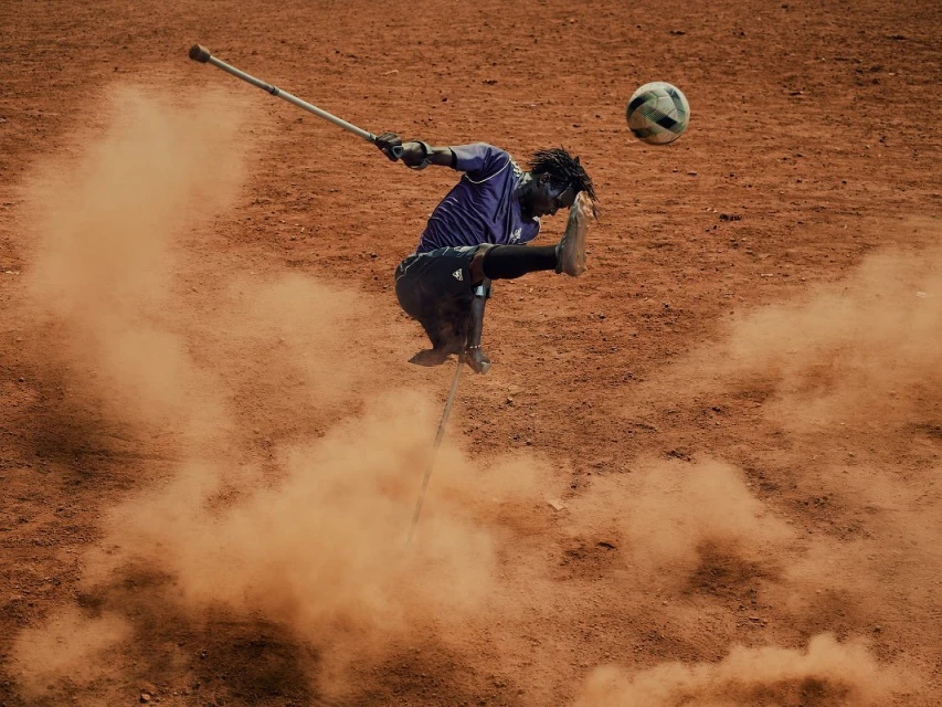 A man with an amputated leg uses crutches to kick a soccer ball in the air on a dusty, reddish-brown surface outdoors on a sunny day.
