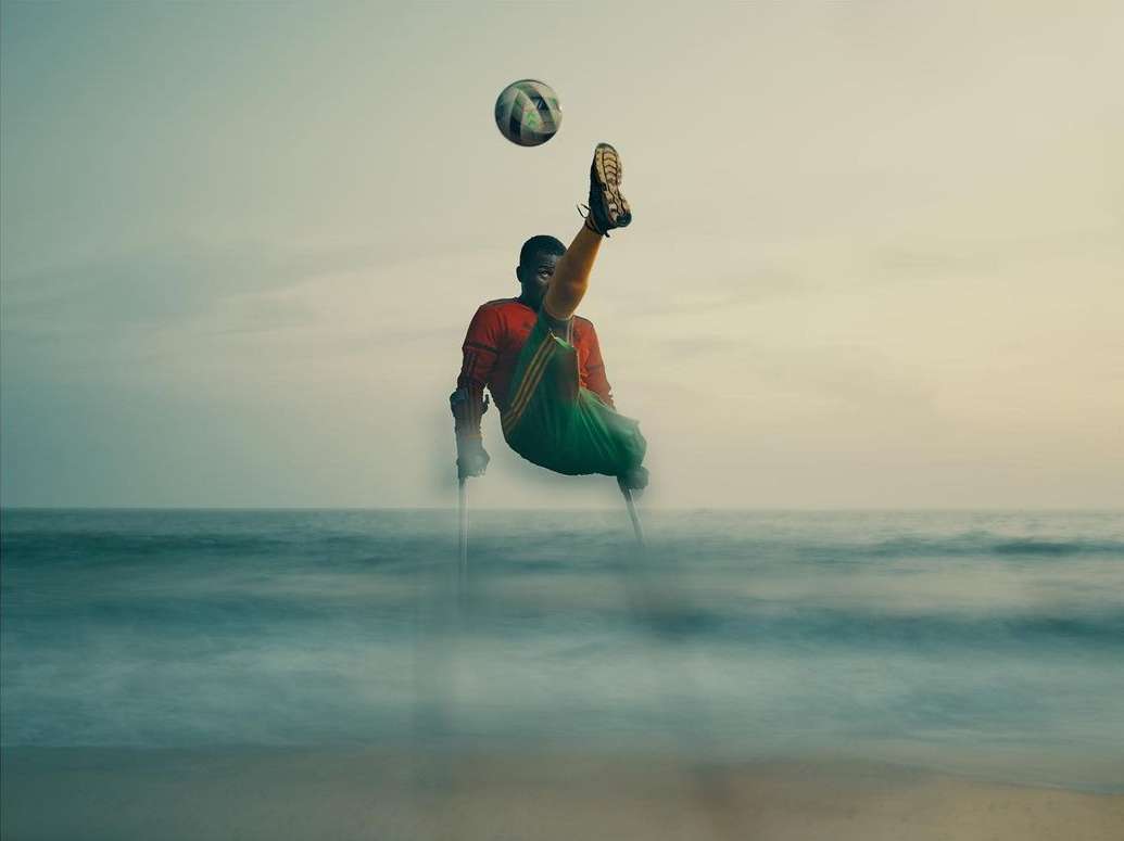 Man with physical disability kicks soccer ball on the beach at sunset. He is using forearm crutches for support and wearing green uniform.