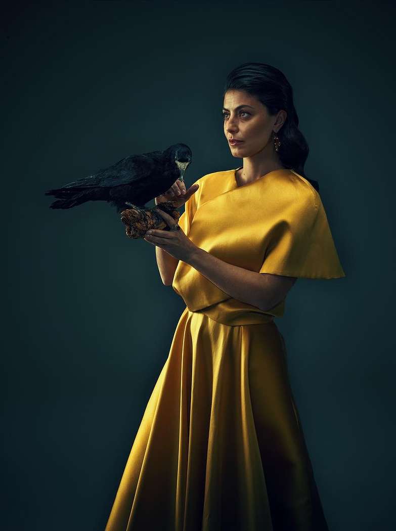 Elegant woman in golden dress holds black raven in studio setting, creating a striking portrait with dark background, eye-level view.