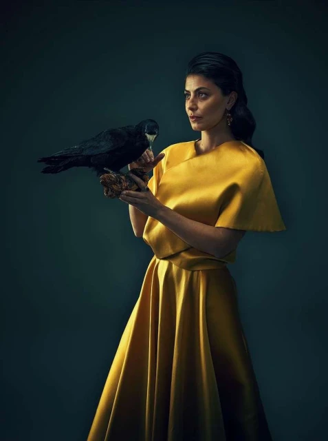 Elegant woman in golden dress holds black raven in studio setting, creating a striking portrait with dark background, eye-level view.