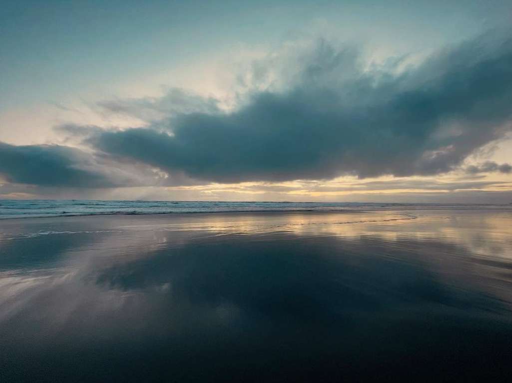 Beach reflecting the twilight sky with dark clouds. Low waves breaking on the shore. Golden light glowing near the horizon.