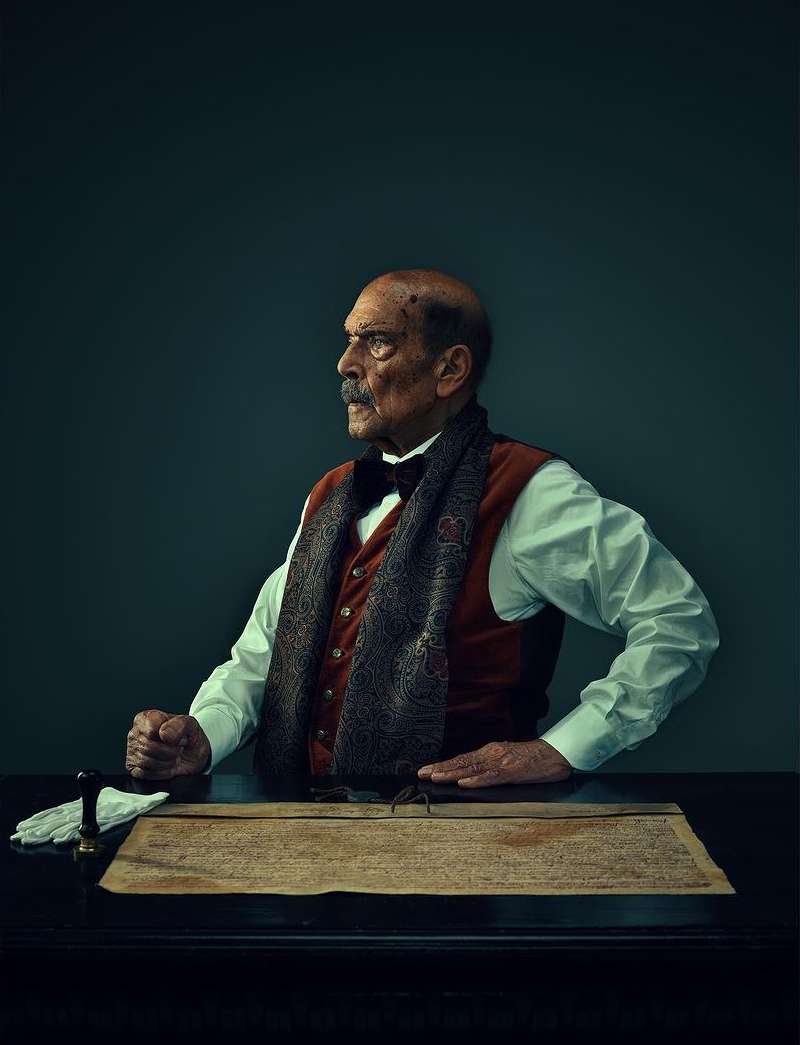 Historical portrait of an elderly man with a mustache wearing a waistcoat and scarf sitting at a table with a document and gloves.