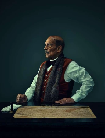 Historical portrait of an elderly man with a mustache wearing a waistcoat and scarf sitting at a table with a document and gloves.